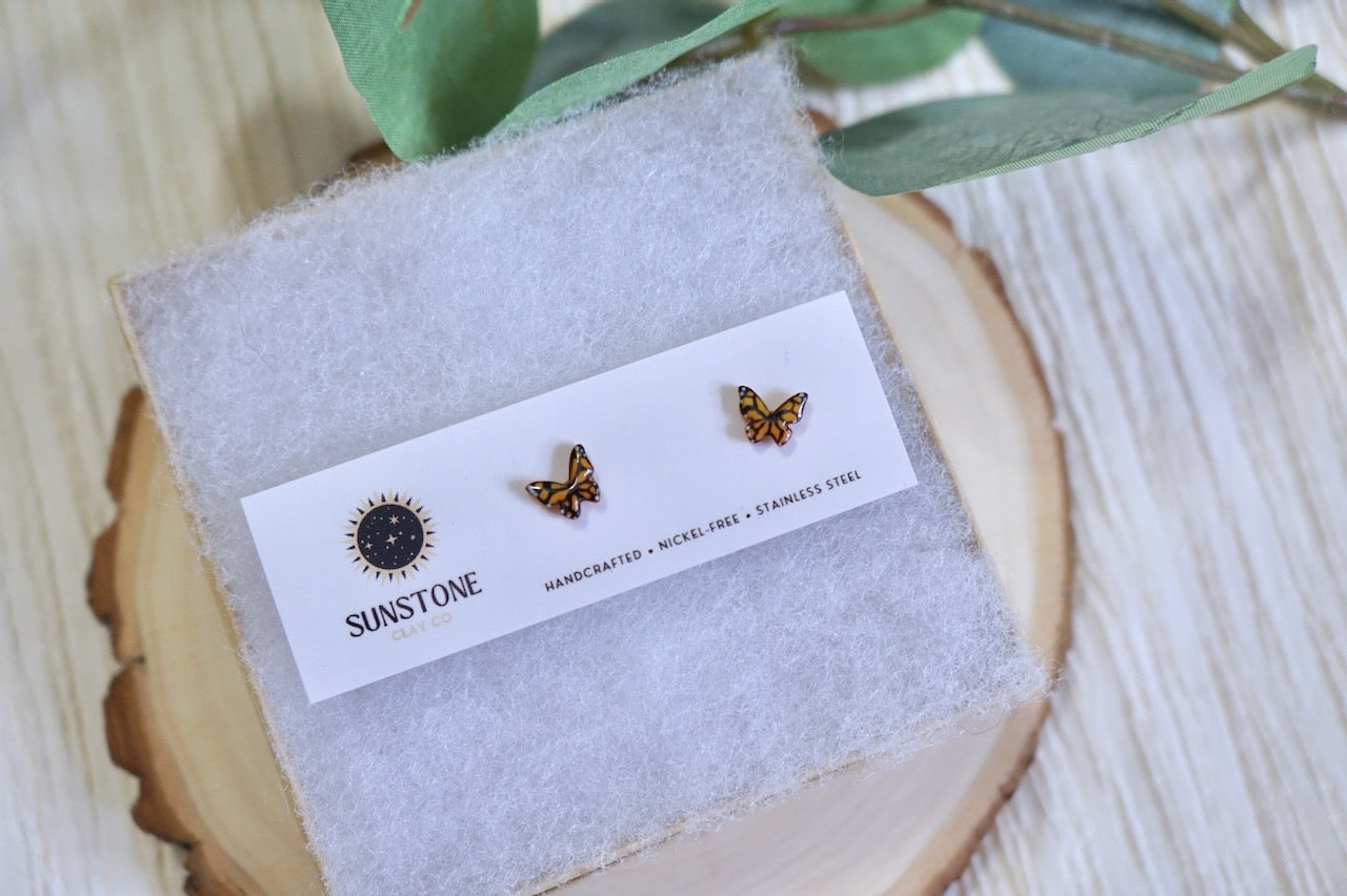 Butterfly-shaped earrings on a display card with 'Sunstone' branding, placed on a wooden surface with green leaves.