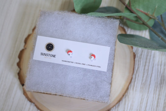 Handcrafted polymer clay mushroom stud earrings by Sunstone Clay Co. on display card, red and white toadstools with stainless steel posts.