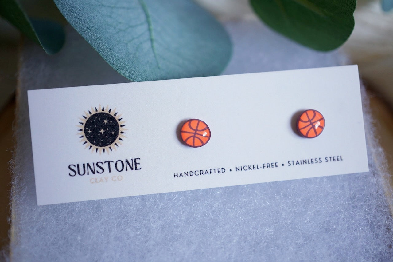Handcrafted basketball polymer clay stud earrings in bright orange, displayed on a Sunstone Clay Co. jewelry card with soft wool padding and greenery styling.