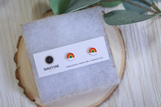 Handcrafted rainbow stud earrings with white cloud base by Sunstone Clay Co., displayed on jewelry card with wood background.