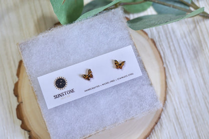 Butterfly-shaped earrings on a display card with 'Sunstone' branding, placed on a wooden surface with green leaves.