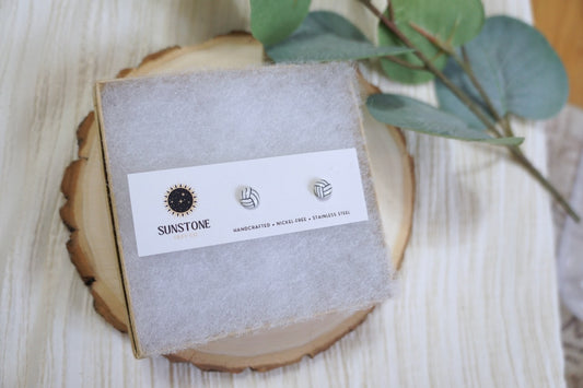 Handcrafted polymer clay volleyball stud earrings displayed on a Sunstone Clay Co. earring card with greenery in the background.