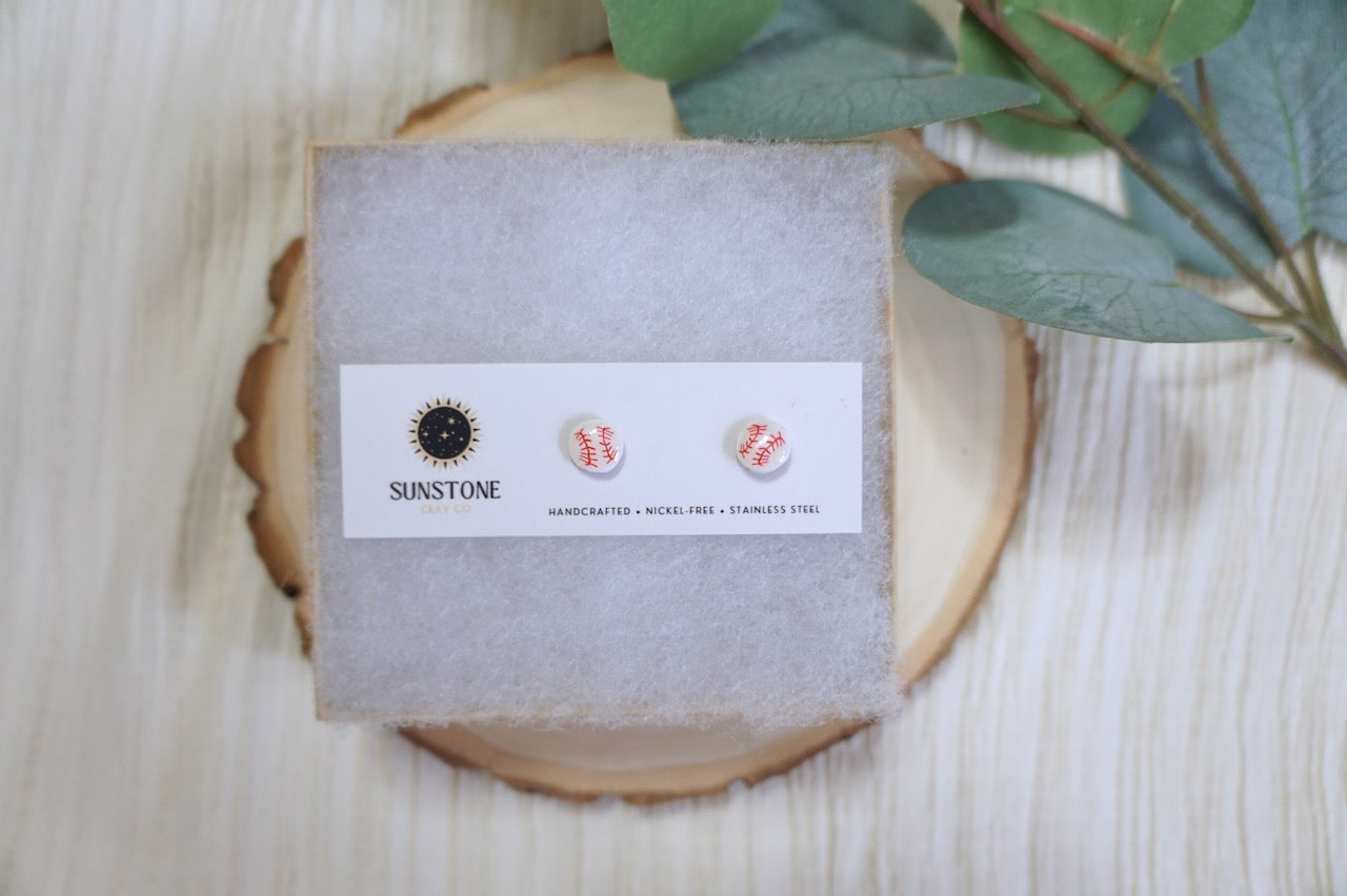 Handcrafted baseball stud earrings with red stitching on a Sunstone Clay Co. earring card displayed on a wood slice background.