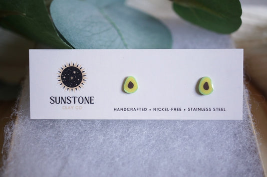 Avocado earrings displayed on a card with a wooden coaster and green leaves in the background