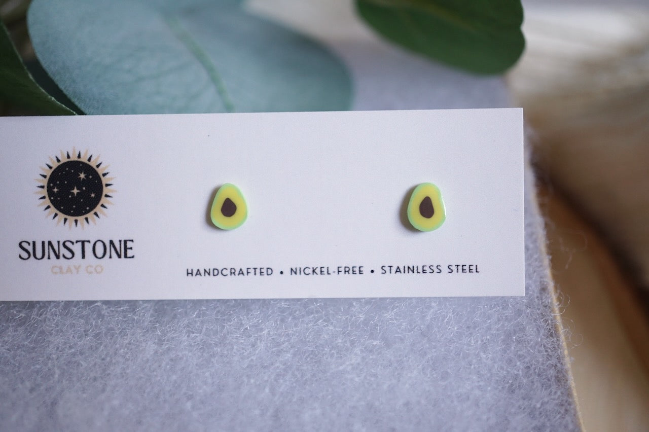 Avocado slice earrings displayed on a card with a wooden coaster and green leaves in the background
