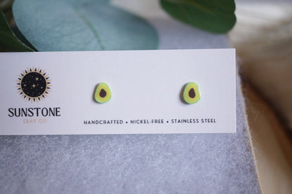 Avocado slice earrings displayed on a card with a wooden coaster and green leaves in the background