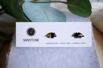 Handcrafted miniature triceratops stud earrings with gold-leaf accents, available in black or gray, displayed on a Sunstone Clay Co jewelry card.