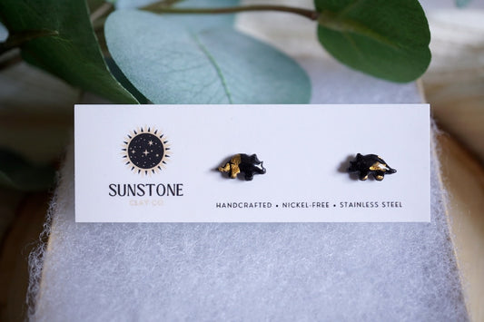 Handcrafted miniature triceratops stud earrings with gold-leaf accents, available in black or gray, displayed on a Sunstone Clay Co jewelry card.