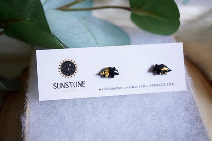 Handcrafted miniature triceratops stud earrings with gold-leaf accents, available in black or gray, displayed on a Sunstone Clay Co jewelry card.