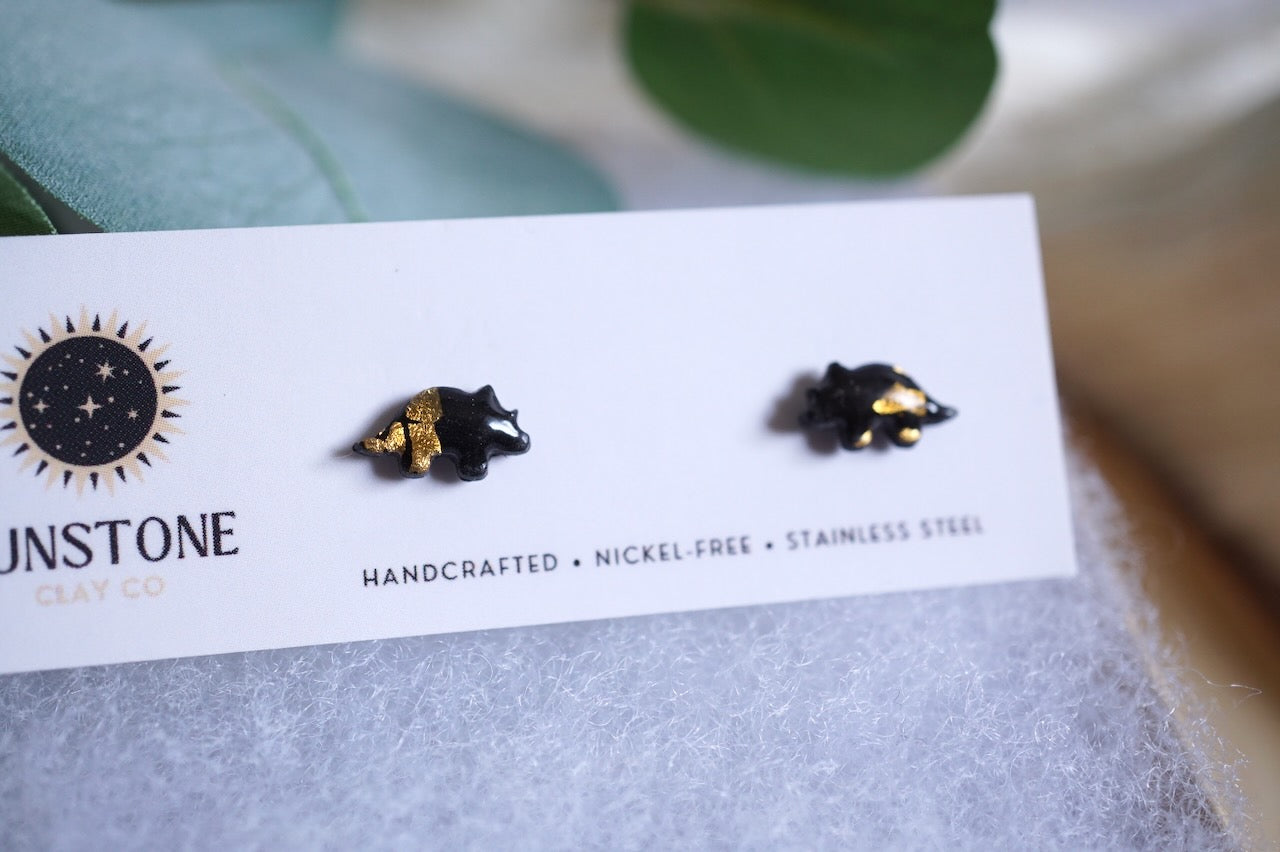 Miniature black and gold triceratops polymer clay studs shown on a branded jewelry card with soft felt backing.