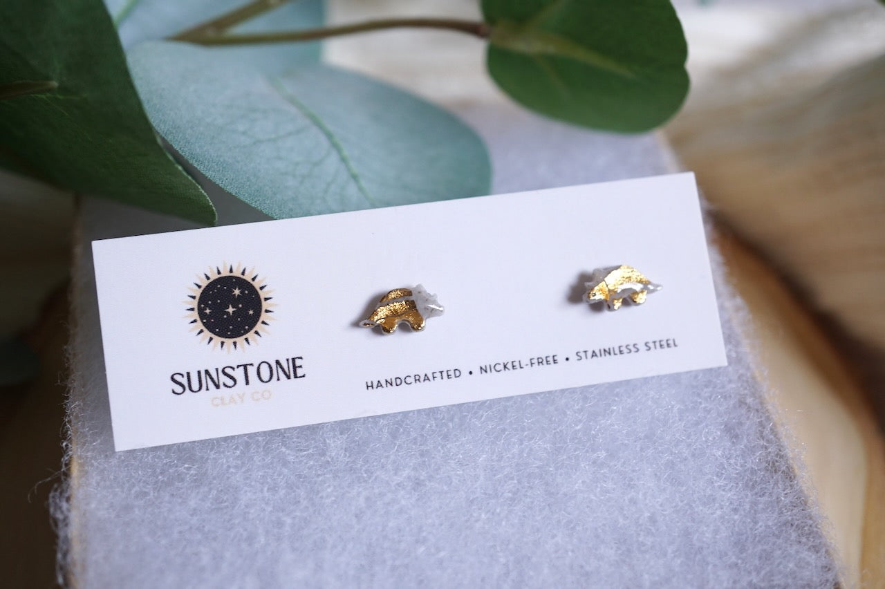 Handcrafted gray triceratops stud earrings with gold-leaf accents on stainless steel posts, displayed on a Sunstone Clay Co jewelry card.