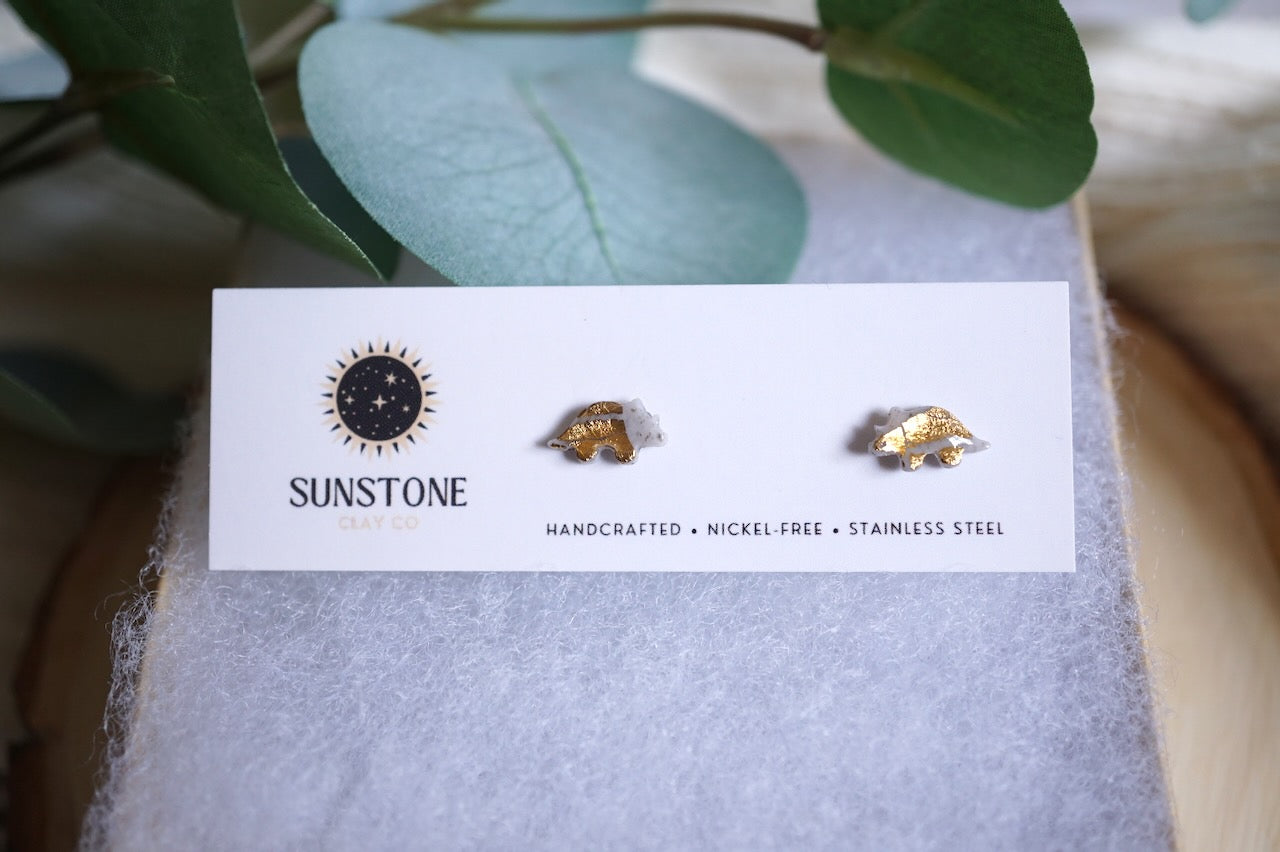 Handcrafted gray triceratops stud earrings with gold-leaf accents on stainless steel posts, displayed on a Sunstone Clay Co jewelry card.