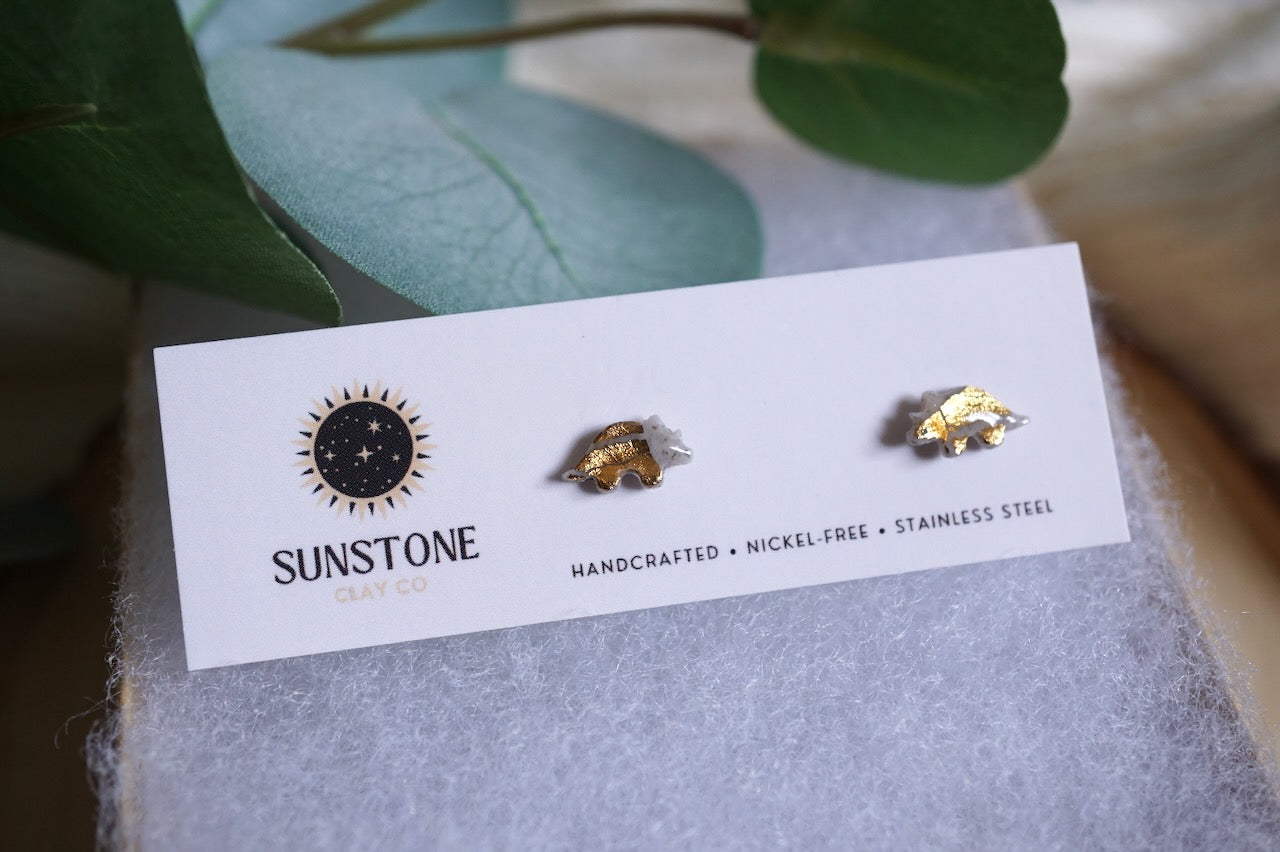 Handcrafted gray triceratops stud earrings with gold-leaf accents on stainless steel posts, displayed on a Sunstone Clay Co jewelry card.