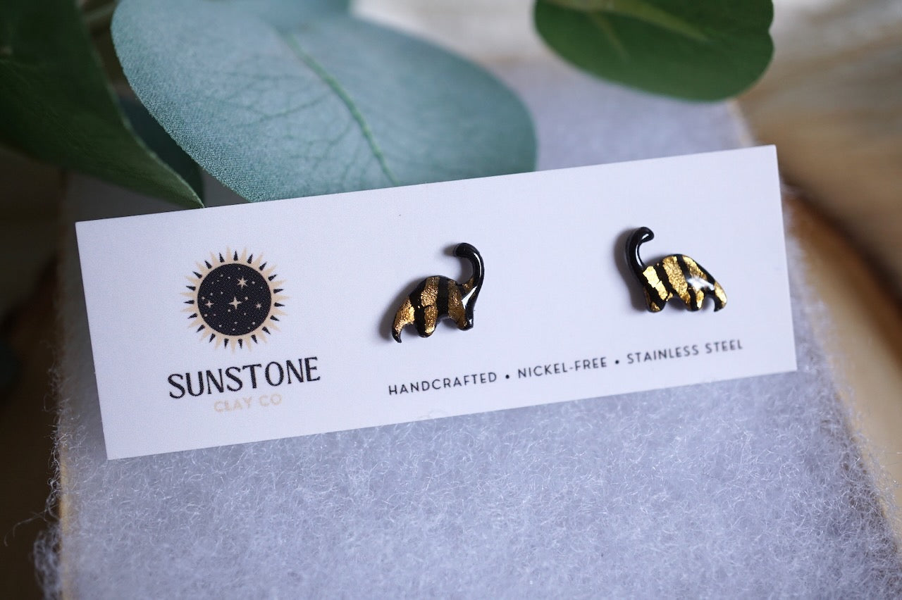 Pair of black and gold brontosaurus earrings on a Sunstone branded card with green leaves in the background.