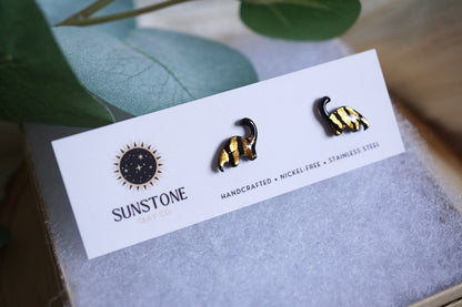 Pair of brontosaurus earrings on a Sunstone branded card with green leaves in the background.