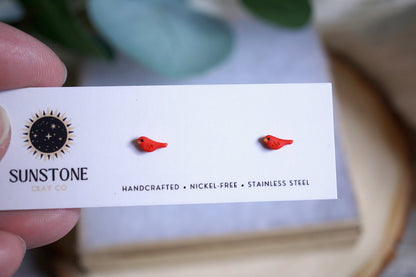 Handcrafted micro red bird stud earrings displayed on Sunstone Clay Co. card.