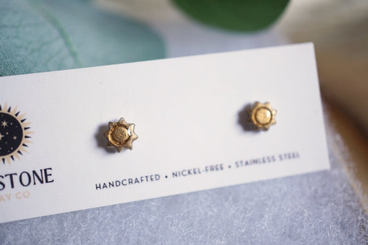 Gold earrings on a white card with 'Stone' branding, displayed on a textured surface.