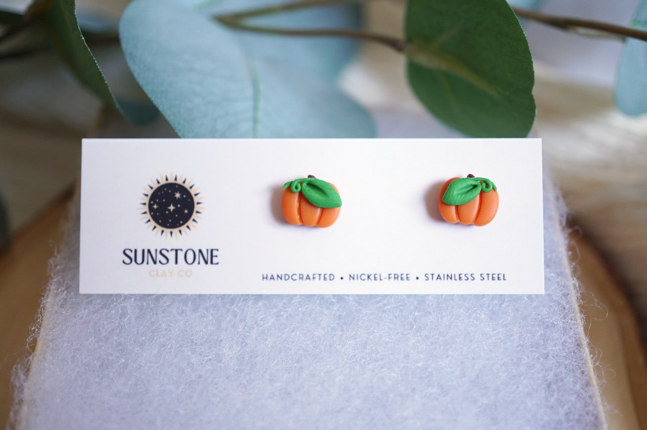 Small orange pumpkin stud earrings with green leaf detail on jewelry backing card