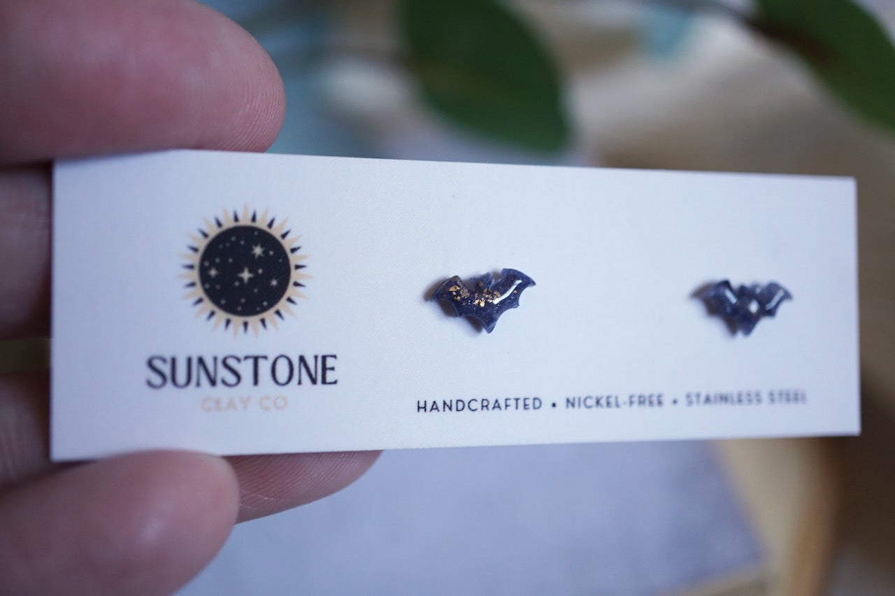 Bat-shaped earrings on a Sunstone Clay Co. card held by a hand.