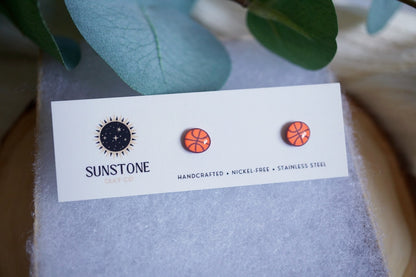 Handcrafted basketball polymer clay stud earrings in bright orange, displayed on a Sunstone Clay Co. jewelry card with soft wool padding and greenery styling.