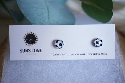 Handcrafted polymer clay soccer ball stud earrings displayed on a Sunstone Clay Co. card with greenery in the background