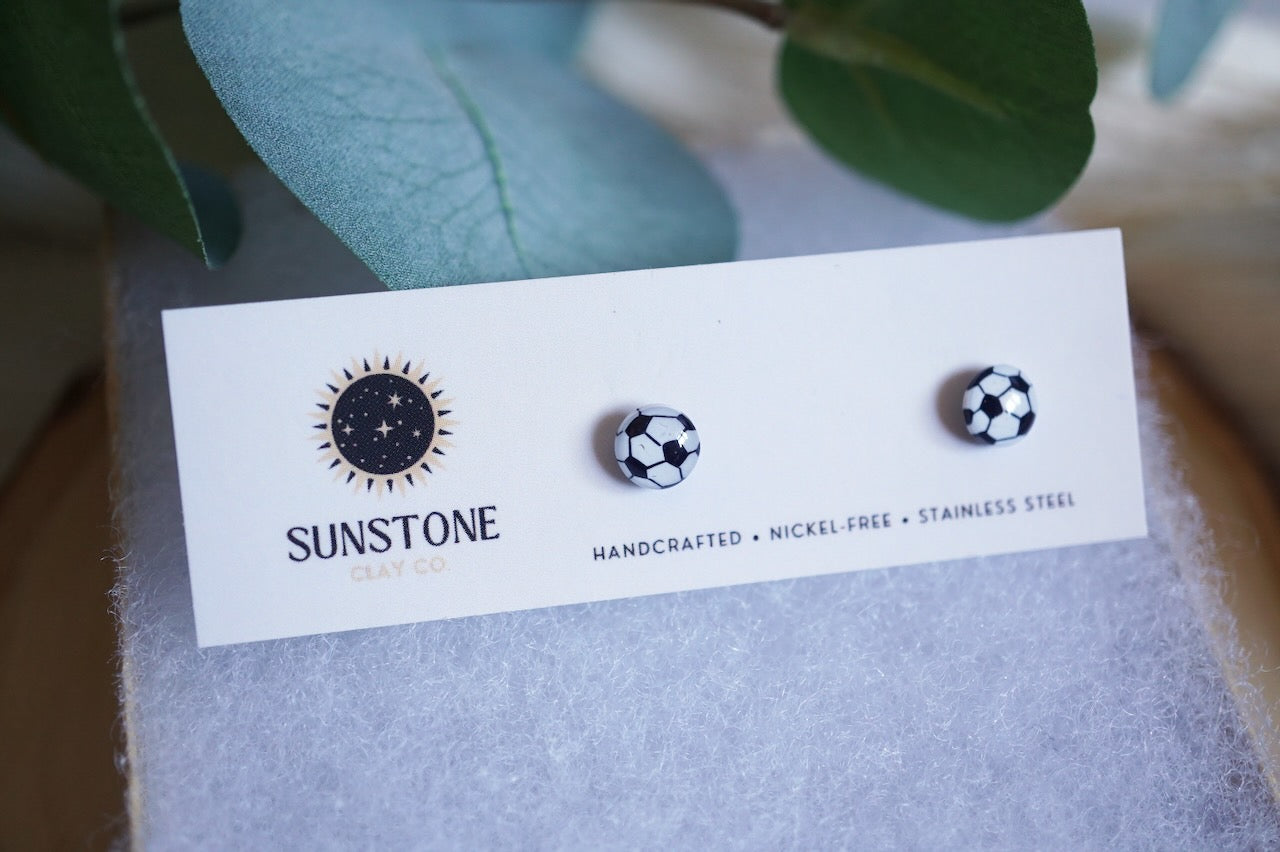 Handcrafted polymer clay soccer ball stud earrings displayed on a Sunstone Clay Co. card with greenery in the background