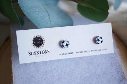 Handcrafted polymer clay soccer ball stud earrings displayed on a Sunstone Clay Co. card with greenery in the background