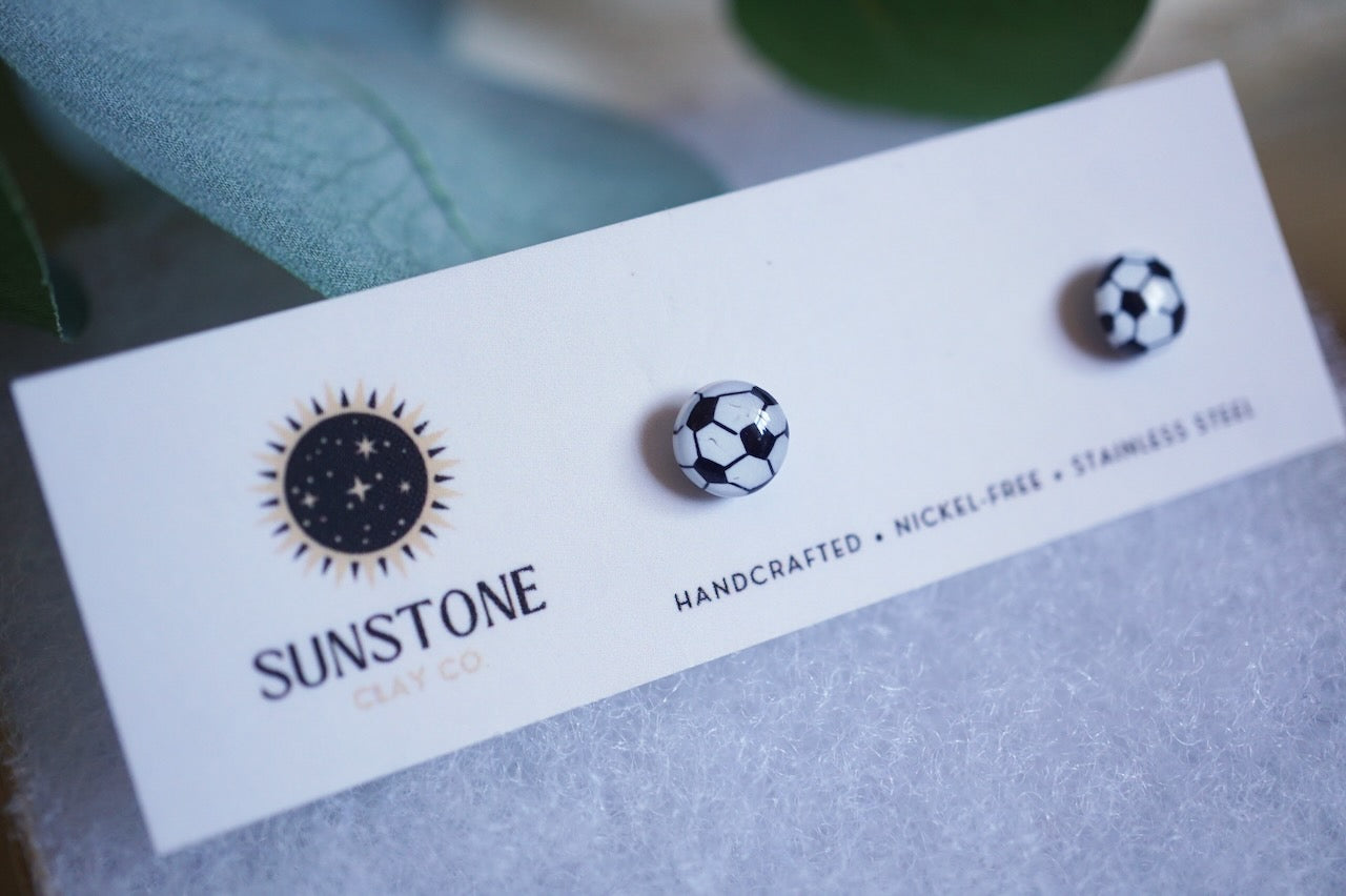 Handcrafted polymer clay soccer ball stud earrings displayed on a Sunstone Clay Co. card with greenery in the background
