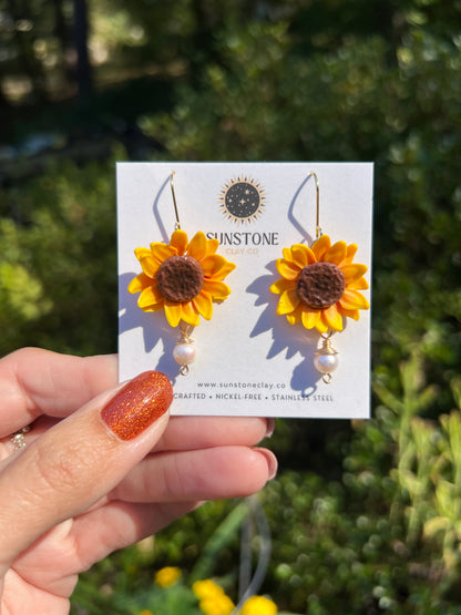 Handmade yellow polymer clay sunflower dangle earrings with textured brown centers, genuine pearl drops, and 18K gold-plated hooks by Sunstone Clay Co.
