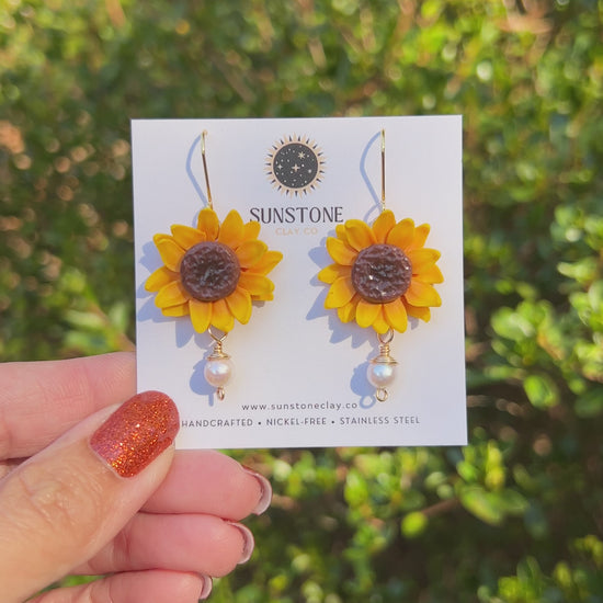 A video of handmade yellow polymer clay sunflower dangle earrings with textured brown centers, genuine pearl drops, and 18K gold-plated hooks by Sunstone Clay Co.