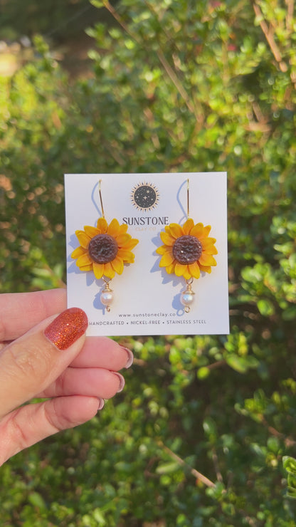 A video of handmade yellow polymer clay sunflower dangle earrings with textured brown centers, genuine pearl drops, and 18K gold-plated hooks by Sunstone Clay Co.